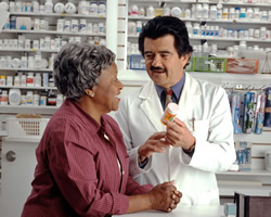 Photograph of a pharmacist with a prescription bottle in his hand talking to an elderly lady.